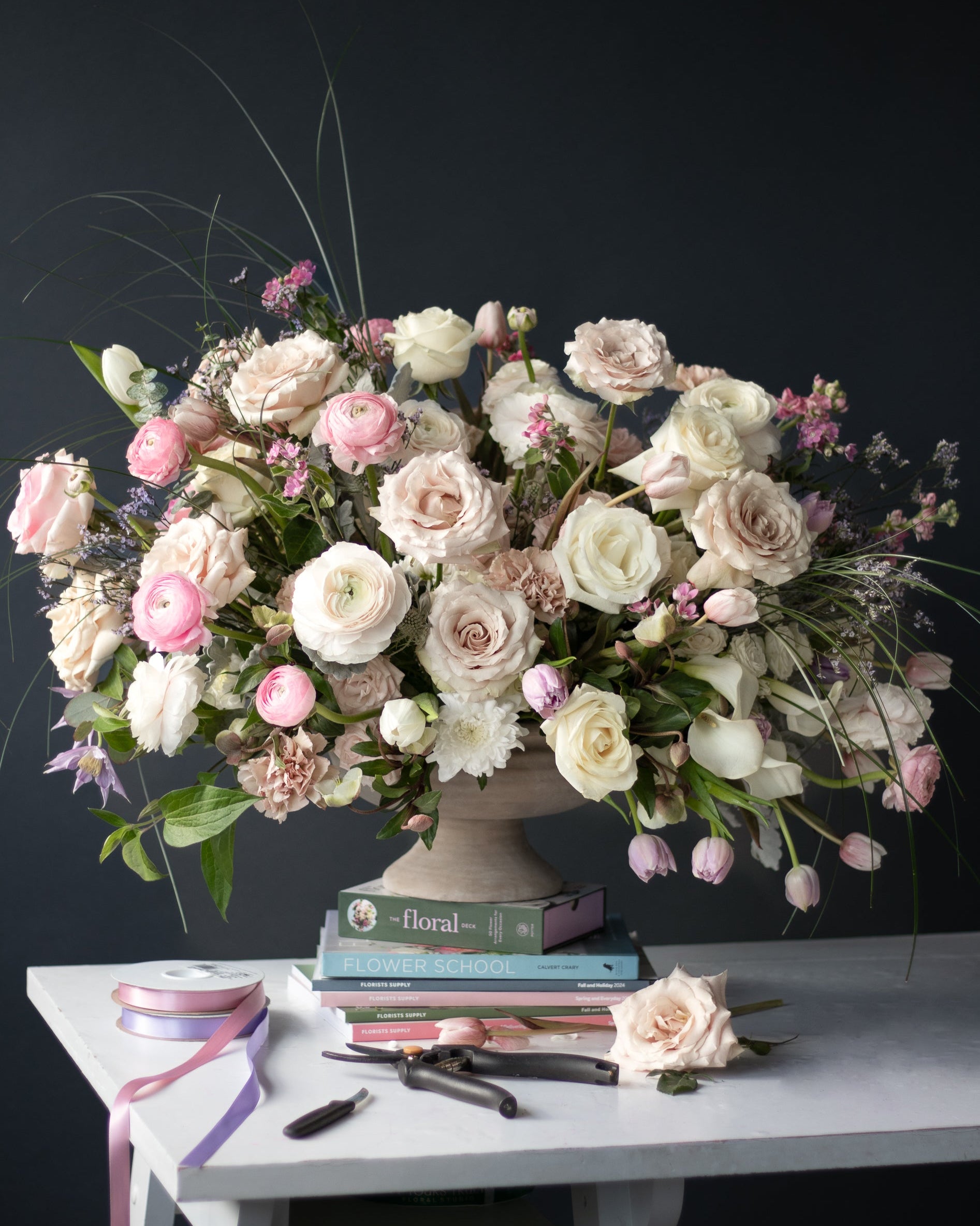 Large floral arrangement in a ceramic compote  on a table with books and tools against a dark background